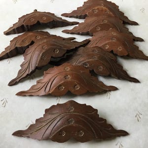 May include: A collection of nine intricately carved wooden leaf-shaped decorative elements. Each piece is a rich brown colour, featuring detailed veining and small floral accents. The leaves are arranged on a patterned surface.