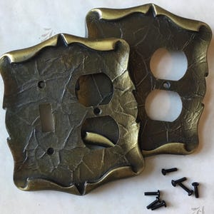 May include: Two decorative, antique-style wall plates. One plate has a switch opening, the other has two outlet openings. Both are bronze-coloured with a textured finish and ornate edges. Small black screws are included.