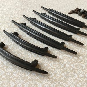 May include: Set of six dark brown, curved drawer pulls with a central handle and two mounting posts. A small pile of screws is also present. The pulls are arranged on a light-colored, patterned surface.