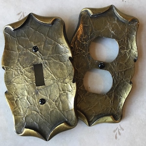 May include: Two decorative wall plates with a textured, metallic gold finish. One plate has a rectangular switch opening and the other has two circular outlet openings. Both plates have black screws and ornate edges.