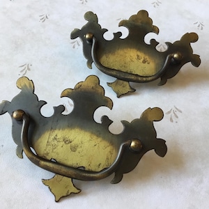 May include: Two ornate brass drawer pulls with a decorative, scroll-like design. The pulls have a slightly aged, antique finish.