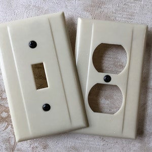 Puede incluir: Dos tapas de tomacorriente de plástico de color crema. Una tapa tiene un interruptor de luz simple y la otra tiene una toma de corriente doble.