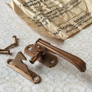 Peut inclure: Un ensemble de loquet de meuble en laiton antique avec des vis et une étiquette en papier. L'ensemble comprend une poignée, un loquet et plusieurs vis. L'étiquette indique "NATIONAL BRASS CO. GRAND RAPIDS, MICHIGAN."