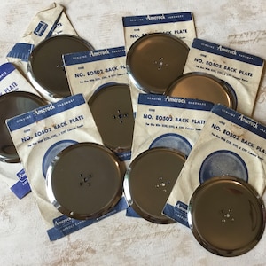 Peut inclure: Gros plan sur huit plaques arrière en métal argenté avec des trous pour vis. Les plaques sont étiquetées "Genuine Amerock Hardware" et "No. E0502 Back Plate".