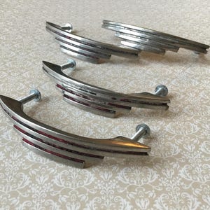 Könnte beinhalten: Vier Vintage-Schrankgriffe in Silberoptik mit einem stromlinienförmigen Art-Déco-Design. Jeder Griff hat eine gebogene Form mit horizontalen Rillen und einem roten Akzent. Die Griffe sind mit Silberschrauben befestigt.