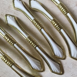 Op de afbeelding: Zes witte en gouden kastknoppen met een decoratief schelpontwerp. De knoppen zijn gemaakt van metaal en hebben een gepolijste afwerking.