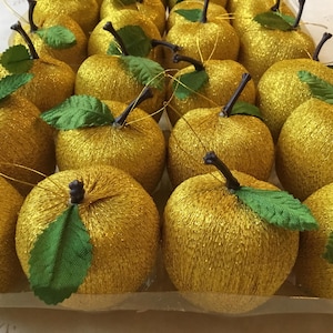 Könnte beinhalten: Eine Ansammlung goldener Apfelornamente mit grünen Stoffblättern und dunkelbraunen Stielen. Die Äpfel sind in Reihen angeordnet und bilden eine festliche Präsentation. Diese dekorativen Ornamente sind ideal für die saisonale Dekoration.