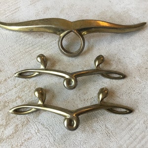 May include: Three gold-toned metal cabinet pulls. The top pull is a long, curved shape with a ring in the centre. The two bottom pulls are smaller, curved shapes with a rounded knob on each end.