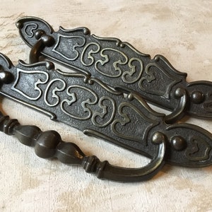 May include: Two antique-style metal drawer pulls with ornate designs. The pulls are a dark bronze color with a textured finish. Each pull has a loop handle and a decorative plate with a floral pattern.