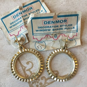 Puede incluir: Dos tiradores de cortina de ventana de metal dorado con un diseño circular y un borde decorativo. Los tiradores están unidos a un tornillo. Los tiradores están etiquetados con "DENMOR DECORATOR STYLED WINDOW SHADE PULLS" y "STYLE 55".