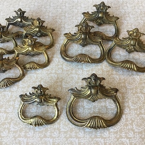 May include: A collection of antique brass drawer pulls. The pulls have a decorative design with a crown-like top and a curved handle. The metal has a vintage, aged appearance, with a mix of gold and darker tones.