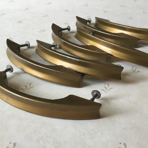 May include: Six vintage gold-coloured drawer pulls. Each pull has a curved design with a pointed end and a screw for mounting. The pulls are arranged on a patterned surface, showing their aged appearance.