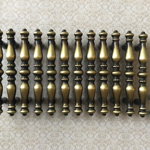 May include: Twelve antique-style brass cabinet pulls with an ornate design. Each pull has a central bulbous shape with detailed accents and a dark finish. A small pile of screws is visible.
