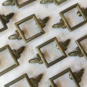 May include: A collection of ten antique brass cabinet knobs with ornate, decorative, scroll-like details. Each knob features a square frame with a round opening and a decorative scroll on each side.