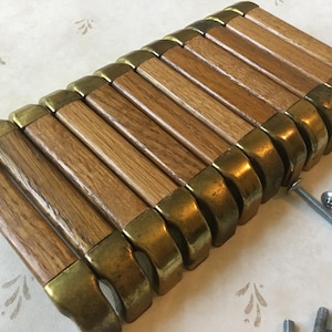 以下が含まれることがあります： 真鍮色の金属アクセントが付いた木製キャビネットプルセット。プルは長方形で、明るい茶色の木目仕上げです。取り付け用の銀色のネジがいくつか見えます。