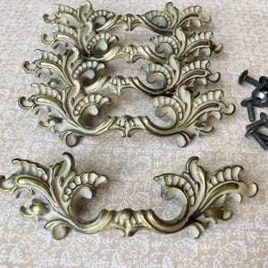 May include: Ornate, vintage-style drawer pulls with a cream and gold finish. The decorative hardware features a scrolling leaf design. Includes several dark screws for installation. A set of cabinet handles.