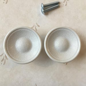 Op de afbeelding: Twee witte keramische kastknoppen met een getextureerd oppervlak. De knoppen hebben een licht verhoogd midden en een afgeronde rand.