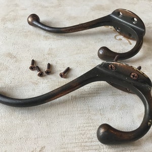 May include: Two antique bronze coat hooks with rounded ends and a decorative back plate. The hooks are mounted on a white surface with four screws.