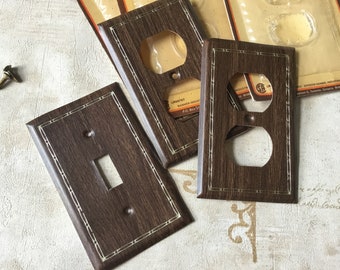 May include: Three vintage brown plastic wall plates with a woodgrain pattern and white trim. One plate has a single light switch, one has a double outlet, and one has a single outlet. The plates are labeled "Lakstrop Banner Industries Inc."