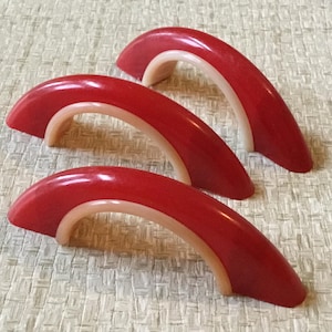 May include: Three red and cream colored plastic cabinet door pulls in the shape of an arch.