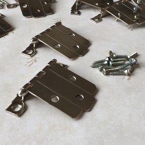 May include: A close-up of ten silver cabinet hinges with a pile of screws. The hinges are made of metal and have three leaves each.