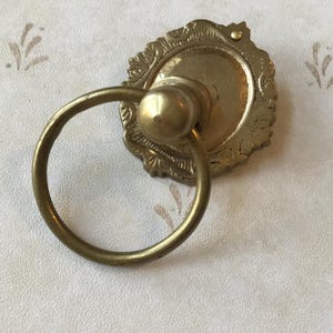 May include: A gold-toned metal ring pull with an ornate, decorative base. The ring pull is attached to a round, flat base with intricate details.