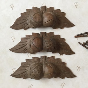 Puede incluir: Tres decoraciones de madera tallada con forma de bellotas y hojas, con tornillos. Las bellotas están partidas, con tallas detalladas de hojas. La madera tiene un acabado marrón oscuro, y los tornillos son una mezcla de plata y óxido.