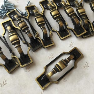 May include: Ten antique brass cabinet drawer pulls with a black finish. Each pull has a decorative design with a raised centre.
