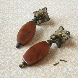 Op de afbeelding: Twee antieke kastgrepen met een warmbruin, ovaalvormig middenstuk en sierlijke, goudkleurige metalen accenten. De grepen worden vergezeld door twee kleine schroeven. De achtergrond is een crèmekleurige stof met een bloemmotief. Klaar voor installatie.