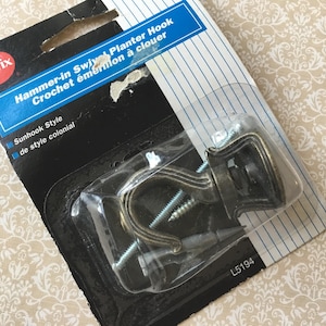 May include: A package containing a bronze-colored hammer-in swivel planter hook. The packaging includes the text "Selfix Hammer-in Swivel Planter Hook Crochet émerion à clouer" and "Sunhook Style de style colonial". The package also contains screws.