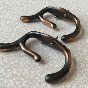 May include: Two antique bronze-colored metal hooks with a curved design. The hooks have a dark brown finish with copper-colored accents and are mounted with screws. These decorative hooks are suitable for hanging items.