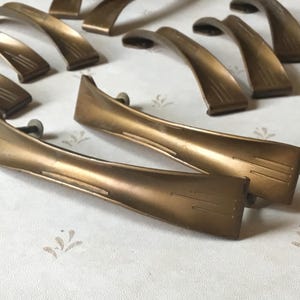 Op de afbeelding: Een verzameling vintage kastgrepen in bronskleur. De grepen hebben een gebogen, gestroomlijnd ontwerp met subtiele lineaire details. De hardware is gerangschikt op een wit oppervlak met een bloemenpatroon, voor interieurdecoratie.