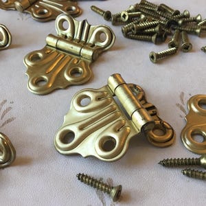 Puede incluir: Un juego de seis bisagras de mariposa doradas adornadas con un diseño decorativo. Las bisagras van acompañadas de un montón de tornillos.