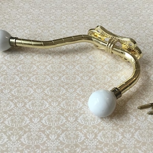 May include: A decorative gold-colored metal wall hook with two white ceramic ball ends. The hook has a curved design and ornate detailing. Includes several small gold screws for mounting.