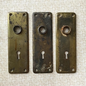 Op de afbeelding: Drie antieke messing deurplaten met sleutelgaten en schroefgaten. De platen hebben een verweerde, verouderde uitstraling met aangetaste plekken. Verschillende kleine messing schroeven liggen in de buurt.