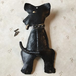 May include: Black metal dog-shaped wall hook with a decorative collar. The hook has a tail that curves upward and two screws are included for mounting. The dog has a textured, painted finish.