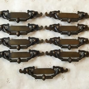 May include: Eight antique brass drawer pulls with ornate designs. Each pull has a rectangular plate with a curved handle.