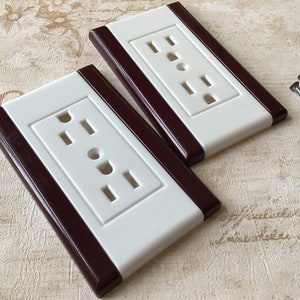 May include: Two white electrical outlet covers with a brown accent stripe. The covers have three slots and a round hole. Two screws are included.