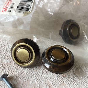 May include: Three brass cabinet knobs with a gold finish and a raised ring design. The knobs are packaged in a clear plastic bag with a label that reads "Amero BP728-AE".