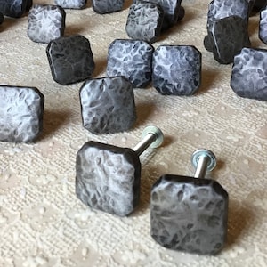 Może przedstawiać: Zbiór 18 kwadratowych gałek do szafek z wykończeniem z młotkowanego metalu. Gałki są ciemnoszare i mają lekko fakturowaną powierzchnię.