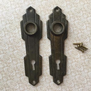 Puede incluir: Dos placas traseras de manija de puerta antiguas de bronce con cerraduras y un diseño decorativo. Las manijas son rectangulares con una abertura circular y una cerradura. Se incluyen varios tornillos de latón.