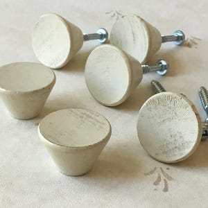 Peut inclure: Six boutons de porte en céramique blanc cassé de forme arrondie légèrement conique. Les boutons ont une finition lisse et mate et sont fixés à des vis argentées.