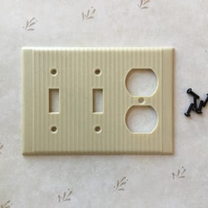 May include: A beige plastic electrical outlet cover with two light switches and a single outlet. The cover has a textured, ribbed surface.