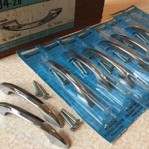 May include: Polished chrome cabinet handles, model T-104-26, with a 10 cm overall length. The handles are packaged in blue plastic and include mounting screws. A vintage Amerock box is visible in the background.