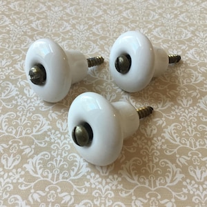 May include: Three white ceramic cabinet knobs with brass hardware. The knobs have a rounded, oval shape and are arranged on a patterned beige surface. These knobs are suitable for furniture restoration or adding a vintage touch to cabinets and drawers.