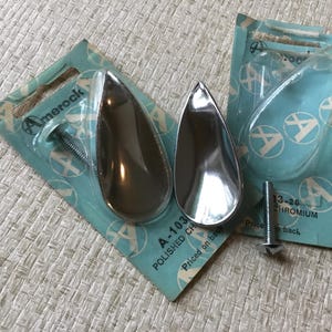 May include: Two chrome teardrop-shaped cabinet knobs with screws, packaged on blue and white cards with the brand name "Amerock" and the product number "A-103". The text "Polished Chrome" is also on the card.