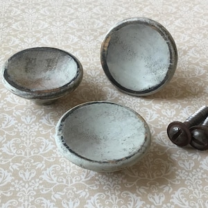 May include: Three distressed white and gray cabinet knobs with a dark rim and two screws. The knobs have a bowl-like shape and are made of a solid material. The screws are dark brown with a flat head.