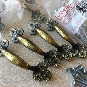 May include: A collection of brass-toned cabinet pulls with a hammered texture. The pulls have a curved handle design and are packaged in clear plastic. A small pile of screws is also visible.