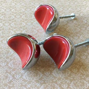 Pode incluir: Três puxadores decorativos para armários com um design vermelho em forma de coração, encapsulados em metal prateado. Os puxadores estão dispostos sobre uma superfície branca suja estampada. Cada puxador tem um parafuso para montagem.