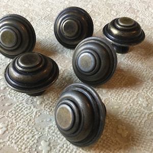 May include: Six antique-style cabinet knobs with a layered, concentric circle design. The knobs are made of metal with a brass finish and have a slightly aged appearance.